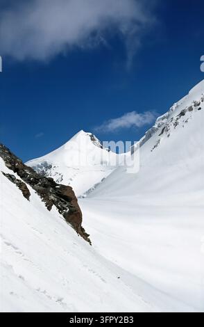 Un sommet majestueux couvert de neige domine la scène sous un ciel bleu éclatant parsemé de nuages. Le paysage présente un terrain rocheux et pri Banque D'Images