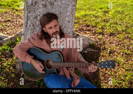 Homme aux cheveux flottants et à la barbe joue de la guitare acoustique tout en chantant, appuyé nonchalamment contre un arbre dans un cadre de parc paisible Banque D'Images