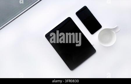 Composition en flat lay d'un bureau propre avec une tablette numérique noire, un téléphone mobile, un stylet et une tasse de café blanche posée sur une surface blanche. Min Banque D'Images
