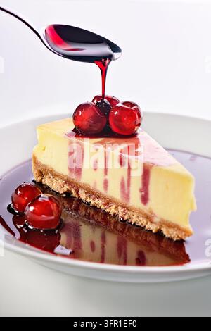 Tranche de cheesecake crémeux avec une croûte de biscuits Graham, garnie de sauce aux cerises rouges, de cerises fraîches et d'une cuillère versant du sirop sur une assiette blanche Banque D'Images