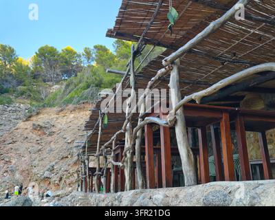 Une terrasse de restaurant en plein air au bord de la mer à Deia, en Espagne, dotée d'une structure de toit rustique en bois et en roseau avec plusieurs tables en bois. Le naturel de la mer Banque D'Images