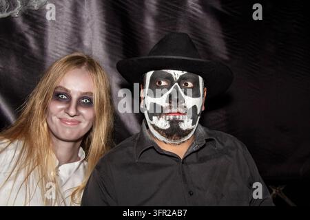 Couple d'Halloween posant devant un fond sombre. Femme fantôme blonde souriante avec des yeux blancs étranges et homme portant un maquillage facial de crâne et un chapeau noir. Banque D'Images
