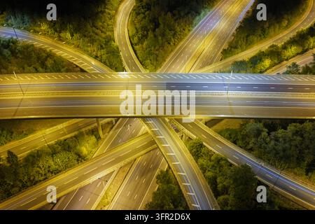 Une vue en contre-plongée montre une intersection autoroutière complexe entourée d'arbres sombres pendant la nuit, tandis que des lampadaires éclairent les routes pavées et Banque D'Images
