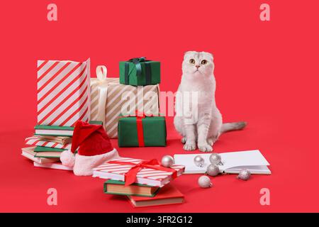 Célébration des fêtes de Noël, chat Scottish Fold assis avec des boîtes-cadeaux emballées et des livres sur un fond rouge pour le marketing de la saison hivernale festive Banque D'Images