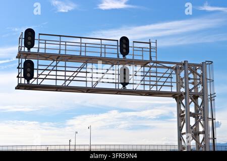 Un grand portique en acier pour signal ferroviaire avec quatre têtes de signal éteintes, des passerelles surélevées et une échelle d'accès se dresse contre un ciel bleu parsemé de légers nuages blancs Banque D'Images