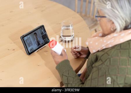 Femme africaine âgée en cardigan tenant une bouteille à bouchon rouge à une table et consultant une tablette, espace libre Banque D'Images
