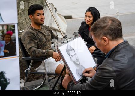 JEUNE HOMME FAISANT POSE POUR SON PORTRAIT, PHOTOGRAPHIE DE RUE REPORTAGE, BARCELONE, ESPAGNE, 2019 : Un jeune homme séduisant portant un haut camouflage pose pour un artiste de rue qui esquisse son portrait caricatural, une femme regardant à ses côtés. INFO : Barcelone est une ville côtière espagnole vibrante, célèbre pour son architecture emblématique, ses plages méditerranéennes, le quartier gothique historique, la riche culture catalane, le football de classe mondiale et une atmosphère métropolitaine animée remplie d'art, de nourriture et de musique. Banque D'Images