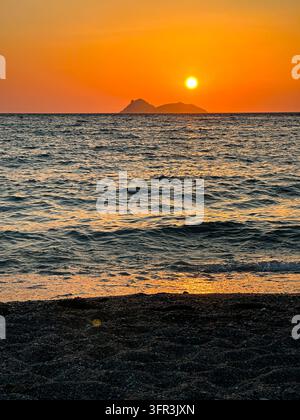Un coucher de soleil orange vif illumine l'océan tandis que le soleil brille au-dessus de la silhouette d'une île lointaine. De douces vagues reflètent une lumière dorée sur des galets sombres Banque D'Images