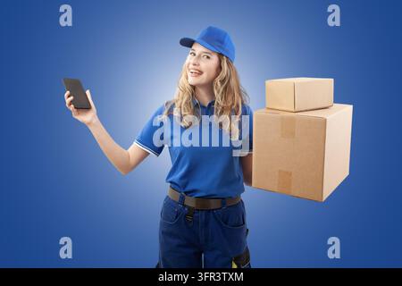 Livraisonneuse joyeuse en uniforme bleu tenant des cartons et présentant un écran de smartphone vierge sur fond bleu. Idéal pour la logistique Banque D'Images