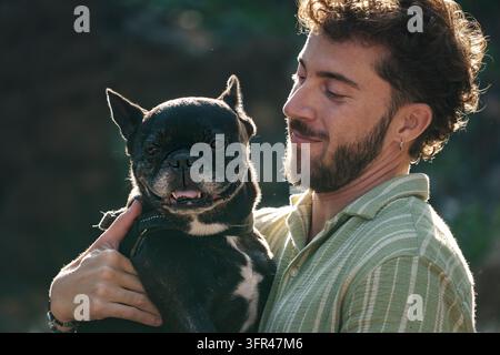 Homme barbu en chemise rayée tenant un bulldog français heureux à l'extérieur, tous deux profitant du soleil et de la nature Banque D'Images