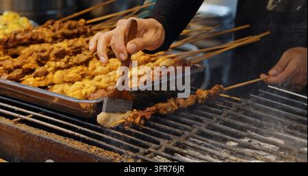 Chef enduisant d'huile des brochettes de viande sur un grill au charbon de bois, marché de rue asiatique traditionnel, processus de cuisine en plein air, documentaire culinaire. Banque D'Images