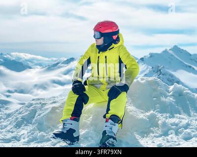 Un adolescent dans un costume de ski de sport lumineux assis sur la neige dans les montagnes Banque D'Images