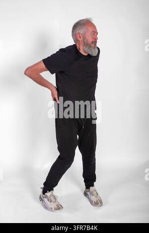 Un cliché en studio en corps entier d'un homme mûr avec une barbe grise et des vêtements de sport noirs, grimacant d'inconfort tout en se tenant le bas du dos avec les deux mains Banque D'Images