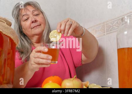 Ancienne femme aux cheveux blancs buvant un verre de thé kombucha fermenté au citron vert naturel Banque D'Images