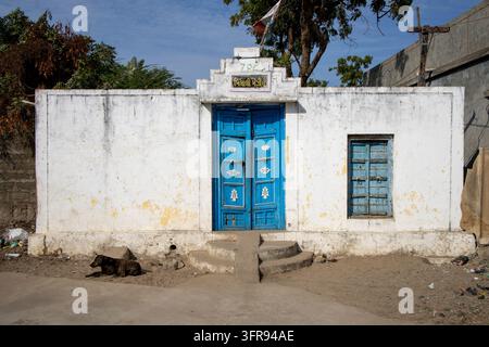 Un chien errant se repose devant une maison blanchie à la chaux avec une porte en bois bleue usée et un panneau au-dessus, Gujarat, Mandvi, Inde Banque D'Images