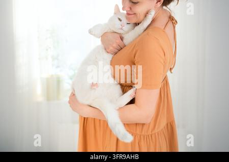 Une femme enceinte tient un chat blanc dans ses bras dans une pièce lumineuse. La mère enceinte qui embrasse son animal de compagnie présente un moment prudent et préoccupé concernant toutes Banque D'Images