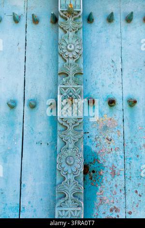 Grossissement d'une porte en bois délavée bleu pâle avec une bande centrale florale sculptée, des clous rouillés et de la peinture écaillée en plusieurs couches en composition verticale Banque D'Images