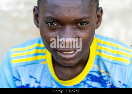 Portrait en gros plan d'un garçon souriant aux yeux chaleureux portant un maillot de football bleu clair avec col et rayures jaunes devant un mur pâle flou en si Banque D'Images