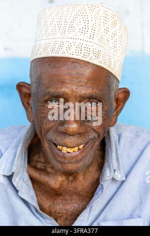 Portrait rapproché d'un homme âgé souriant avec des rides profondes portant une cofia blanche brodée et une chemise bleu clair contre un fond bleu et blanc Banque D'Images