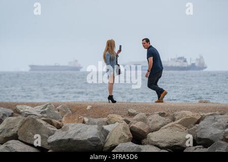 POSE JOUEUSE POUR UNE PHOTO DE PLAGE, REPORTAGE DE PLAYA DE LA BARCELONETA, BARCELONE, ESPAGNE : Une femme en talons hauts tient son téléphone pour photographier un homme qui prend une pose dynamique et ludique pour elle sur un brise-lames rocheux, avec de grands navires visibles sur l'océan derrière eux. INFO : La plage de Barceloneta est la destination balnéaire la plus célèbre et vibrante de Barcelone. Célèbre pour ses sables en pente et sa promenade animée bordée d'arbres, elle allie bains de soleil et sports nautiques avec des bars et restaurants animés en bord de mer, des jetées en béton et des zones d'exercice. Banque D'Images