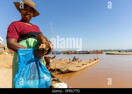 Une femme tient une poule et un sac contenant ses affaires à côté d'une rangée de pirogues amarrées sur la rive, Menabe, Tsiribihina, Madagascar Banque D'Images