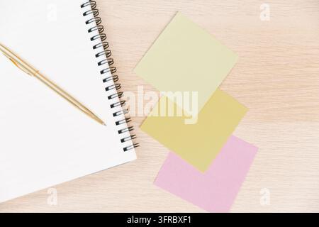 Vue de dessus d'un carnet à spirale ouvert, un stylo doré et des post-it vierges colorés sur un bureau en bois, représentant la planification, les rappels, l'organisation, la production Banque D'Images