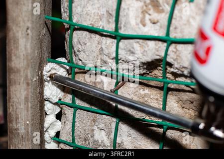 Un gros plan d'un ouvrier du bâtiment utilisant un pistolet métallique pour appliquer une mousse de polyuréthane expansible dans les interstices des murs au-dessus d'un treillis de renfort vert. Profession Banque D'Images