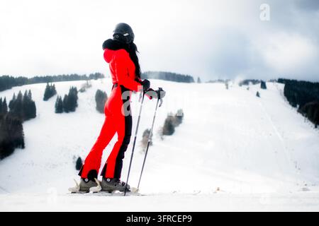Ruzomberok, Slovaquie - 4 janvier 2026 : Skieuse en combinaison de ski rouge se tient avec ses bâtons de ski, regardant les pistes enneigées et les forêts de Malino Brdo Banque D'Images