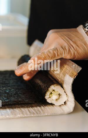 Le chef roule des sushis farcis de riz et de légumes à l'aide d'un tapis en bambou recouvert de plastique. Représente les traditions culinaires japonaises, la nourriture faite à la main et pro Banque D'Images