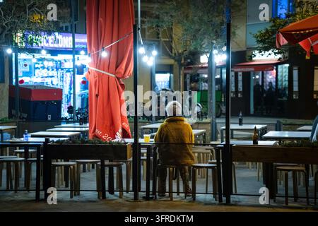 PERSONNE SOLITAIRE DANS UN CAFÉ EN PLEIN AIR DE NUIT, PHOTOGRAPHIE DE RÉPORTAGE DE RUE DE LA VIE NOCTURNE DE BARCELONE, BARCELONE, ESPAGNE : Une personne solitaire portant une veste jaune est assise dos à l'appareil à une table en terrasse d'un café la nuit, une bouteille de bière à côté d'elle. INFO : Barcelone est une ville côtière espagnole-catalane dynamique, réputée pour son architecture emblématique, ses plages méditerranéennes, le quartier gothique historique, sa culture nocturne, le football de classe mondiale et une atmosphère métropolitaine animée remplie d'art, de nourriture et de musique. Banque D'Images