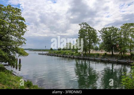 Canal tranquille avec des piliers en bois menant à la vaste étendue du lac de Constance sous un ciel nuageux, créant une scène sereine et pittoresque Banque D'Images