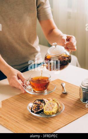 Une femme verse du thé noir chaud d'une théière dans une tasse en verre, consommation de thé fait maison. Espace de copie, mise au point sélective, cadre vertical Banque D'Images
