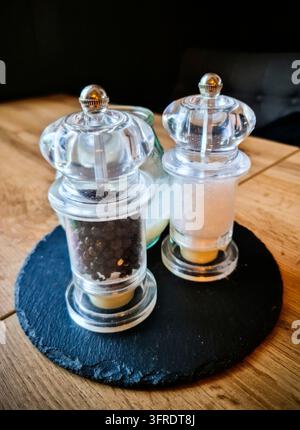 Les moulins à sel et à poivre transparents sont installés sur un dessous de verre en ardoise sombre, ajoutant une touche d'élégance à la table d'un restaurant Banque D'Images