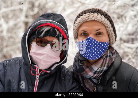 Une femme âgée et sa fille plus jeune vêtue d'hiver, toutes deux portant un masque facial de virus, une neige floue couvrait les arbres en arrière-plan Banque D'Images