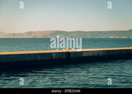 Pier en béton s'étendant sur l'eau bleu profond de la mer avec des montagnes côtières rocheuses à l'horizon sous une lumière chaude du soleil. Mise au point sélective. Banque D'Images