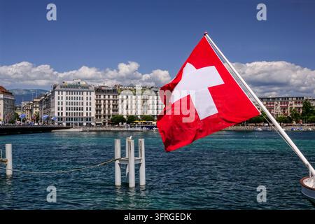 Un drapeau suisse flotte sur l'eau bleue et claire du port devant les grands bâtiments du front de lac à Genève, Suisse, le 17 juillet 2024. Banque D'Images