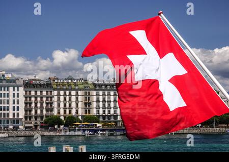 Un drapeau suisse flotte sur l'eau bleue et claire du port devant les grands bâtiments du front de lac à Genève, Suisse, le 17 juillet 2024. Banque D'Images