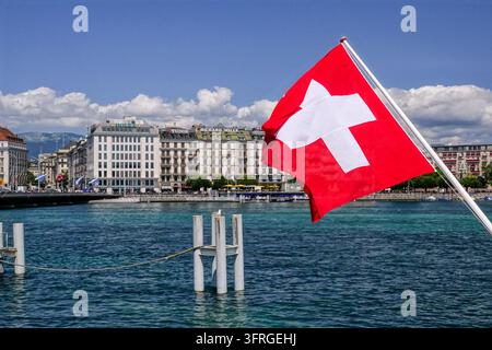 Un drapeau suisse flotte sur l'eau bleue et claire du port devant les grands bâtiments du front de lac à Genève, Suisse, le 17 juillet 2024. Banque D'Images