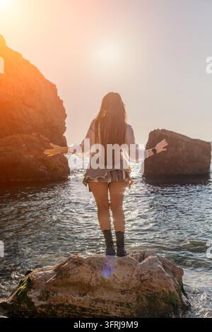 Femme au coucher du soleil sur l'océan, debout sur un rocher les bras écartés vers l'eau pendant l'heure dorée du coucher du soleil pour un sentiment de liberté Banque D'Images