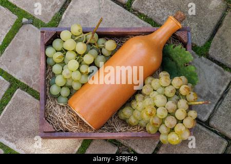 Vue de dessus d'une bouteille en céramique brune posée en diagonale parmi des grappes de raisins verts et des feuilles de vigne dans une caisse en bois sur des pavés gris. Banque D'Images