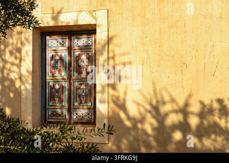Fenêtre marocaine rustique avec volets en bois peints à la main sur un mur de plâtre jaune à Marrakech, baignée d'une lumière chaude du soleil avec des ombres feuillues. Banque D'Images