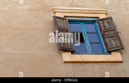 Fenêtre marocaine rustique avec volets en bois peints ouverts sur un mur en adobe ocre texturé, offrant une esthétique architecturale historique et sereine. Banque D'Images