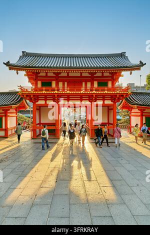 Les touristes traversent la porte ouest shinto vermillon sous le soleil du printemps en fin d'après-midi, projetant de longues ombres, avec Shijo-dori visible au loin Banque D'Images