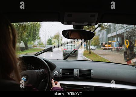 Urdorf, Zurich, Suisse, 10 octobre 2026. Femme conduisant une voiture BMW sur une rue en asphalte mouillée pendant une journée d'automne pluvieuse, vue depuis l'intérieur du tableau de bord du véhicule Banque D'Images