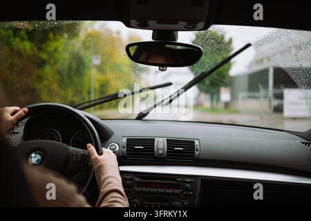Urdorf, Zurich, Suisse, 10 octobre 2026. Les mains d'une femme tenant le volant tout en conduisant une voiture BMW sur une rue de banlieue mouillée pendant un automne pluvieux Banque D'Images