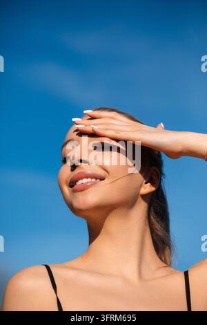 Jolie femme souriant sous un ciel bleu éclatant naturellement. Portrait d’un modèle féminin mettant en valeur une peau saine et radieuse en plein air. Concept de beauté estivale. Banque D'Images