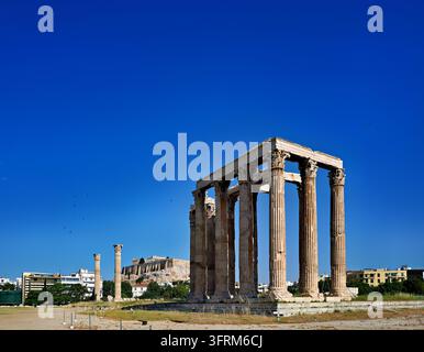 Anciennes colonnes corinthiennes de l'Olympieion sous un ciel bleu clair du matin, avec le Parthénon au sommet de la colline de l'Acropole au loin Banque D'Images