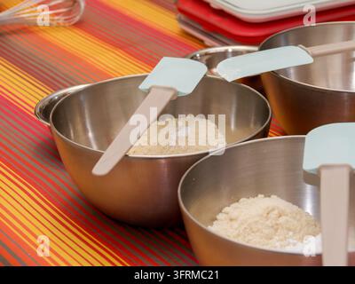 Trois bols à mélanger en métal avec différentes ingrédients secs pour la pâtisserie et des spatules en silicone sur une surface rayée Banque D'Images