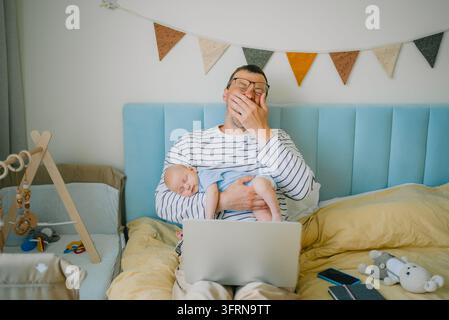 Père fatigué bâillant, tenant un nouveau-né endormi tout en utilisant un ordinateur portable au lit, représentant les défis de concilier parentalité et travail Banque D'Images