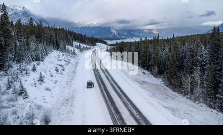 Vue aérienne par drone d'un voyageur en veste orange faisant signe à côté d'un véhicule tout-terrain garé sur une autoroute enneigée près du lac Abraham, en Alberta, Canada. Banque D'Images